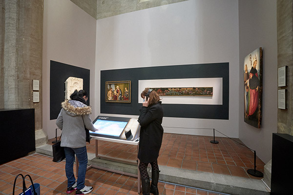 Eglise du musée, devant le tableau "La Chasse", deux personnes utilisent un dispositif de médiation composé d'un grand écran tactile et d'un casque d'écoute.