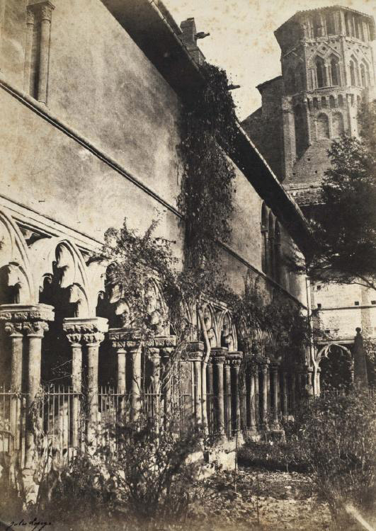 Vue ancienne du grand cloître, datée 1855 : une colonnade avec des grilles, de la végétation envahissante dans le jardin du cloître, le clocher en arrière-plan.
