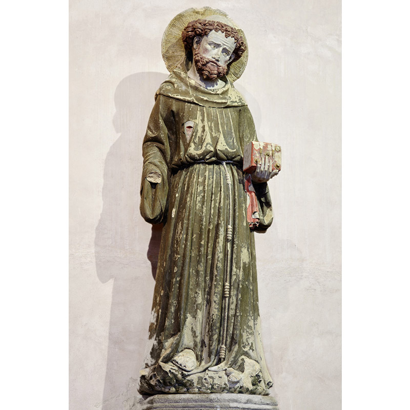 Statue en pierre calcaire polychrome représentant saint François d'Assise, tête penchée et auréolée, barbe et cheveux bouclés. Il tient un livre, sûrement la Bible.